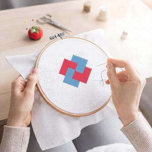 May include: A cross-stitch project in progress, featuring a geometric design in red and blue on white fabric stretched within a wooden embroidery hoop. The scene includes a tomato-shaped pin cushion, scissors, and thread spools.