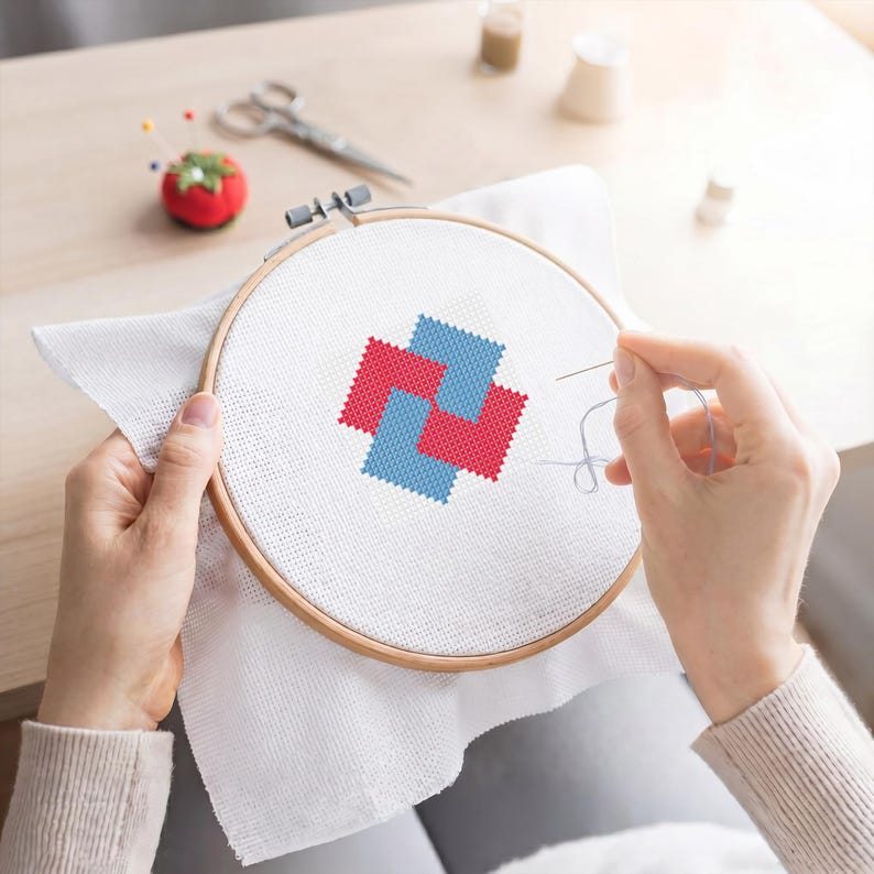 May include: A cross-stitch project in progress, featuring a geometric design in red and blue on white fabric stretched within a wooden embroidery hoop. The scene includes a tomato-shaped pin cushion, scissors, and thread spools.