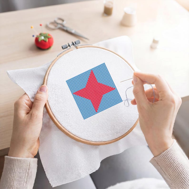 May include: A cross-stitch project in a wooden hoop features a red star on a blue square. The needle is threaded with white thread, and the fabric is stretched taut. A tomato-shaped pin cushion and scissors are in the background.