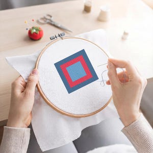 May include: A cross-stitch project in a wooden embroidery hoop. The design features a square within a square, with a light blue center, a red border, and a dark blue outer square. A needle and thread are in use, indicating the project is in progress.