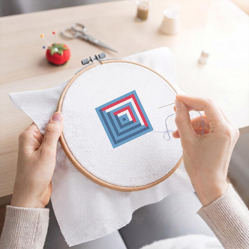 May include: A cross-stitch project in progress, with a geometric design in red and blue within a wooden embroidery hoop. The needle is threaded with light blue thread. A tomato-shaped pin cushion and scissors are in the background.