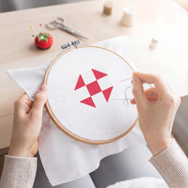 May include: A cross-stitch project in a wooden embroidery hoop. The white fabric features a red and white geometric design. A needle and thread are being used to complete the stitching. Other craft supplies are in the background.