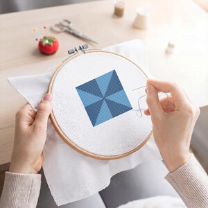 May include: A cross-stitch project in a wooden embroidery hoop. The design features a geometric pattern in shades of blue. A needle and thread are in use, with a red tomato pin cushion and scissors in the background. Crafting and DIY.