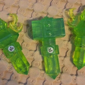 May include: Three translucent green toy figures with yellow accents. Each figure has a geometric design with a logo and a pointed end. The figures are arranged on a textured surface.
