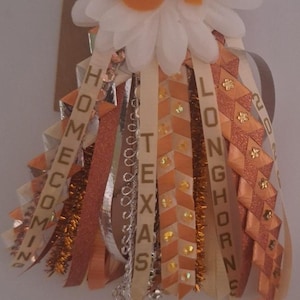 May include: A homecoming mum with a white flower base and orange "UT" letters. Ribbons in orange, white, and gold feature the words "HOMECOMING", "TEXAS", and "LONGHORNS". A yellow tag displays "# 1".
