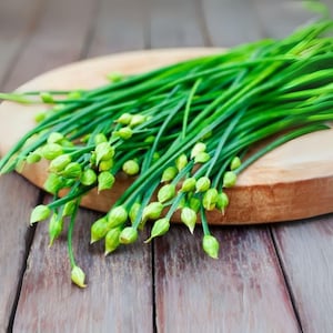 May include: A bundle of fresh green chives with small, unopened flower buds, resting on a round wooden cutting board. The chives are vibrant green, contrasting with the light brown of the board and the rustic wooden background.