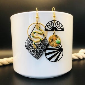 Op de afbeelding: Een paar ongelijke oorbellen met goudkleurige haken. De ene oorbel toont een gouden slang in een cirkel boven een zwarte filigraan ruitvorm. De andere heeft een zwarte waaier, een gouden zonneschijf met een groene steen en een zwarte cirkel.