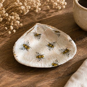 Puede incluir: Un plato de concha blanca decorado con abejas amarillas y negras pintadas a mano. El plato está sobre una superficie de madera, con una taza y flores secas en el fondo. Un artículo decorativo para el hogar.