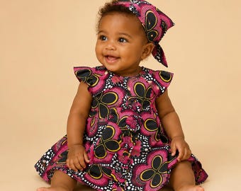Op de afbeelding: Een mouwloze jurk en bijpassende hoofddoek met een levendige, op Afrika geïnspireerde print met gedurfde zwarte, magenta en gele bloemmotieven. De jurk heeft ruche-mouwen en een geplooide rok, terwijl de hoofddoek stevig is vastgeknoopt.