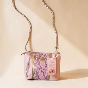 May include: A pink crossbody bag with a silver chain strap. The bag features a snakeskin pattern in shades of pink and gold accents. A tag with the text "Wingie Blossom" hangs from the bag.
