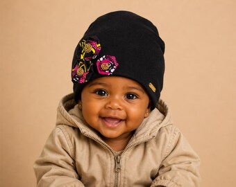 Op de afbeelding: Een zwarte beanie-muts versierd met drie kleurrijke stoffen bloemaccenten. De muts heeft een klein goudkleurig label. De baby draagt een beige jas met capuchon en lacht.