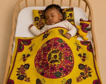 Op de afbeelding: Een slapende baby in een houten wieg met een geel en magenta patroon deken en kussen. Een geel en magenta knuffel ligt naast de baby. De deken en het kussen hebben een bloemen- en vlinderontwerp.
