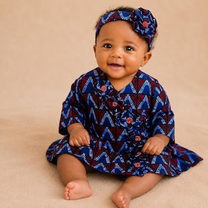 May include: A baby in a blue and red patterned dress and matching headband with a flower. The dress has a V-neck and ruffled detailing. The baby is smiling and sitting on a neutral-coloured surface.
