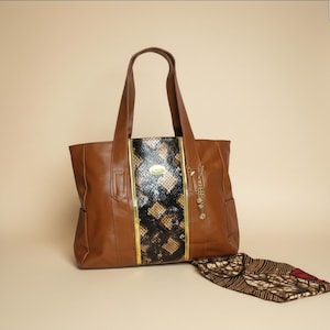 May include: A brown leather tote bag with a snakeskin-patterned center panel and gold accents. The bag has two handles and a small charm. A patterned scarf is next to the bag.