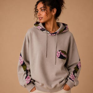 Könnte beinhalten: Ein grauer Kapuzenpullover mit Kordelzug und Kängurutasche. Das Kapuzenfutter, die Tasche und die Ärmel weisen ein Blumenmuster mit rosa und grünen Akzenten auf. Das Model trägt eine beige Hose.