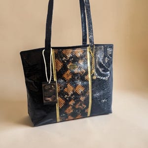 Op de afbeelding: Zwarte tote bag met een slangenprint middenpaneel en gouden rand. De tas heeft twee zwarte handvatten en een kleine gouden ketting. Een label hangt aan het handvat. De tas is gemaakt van een glanzend, getextureerd materiaal.