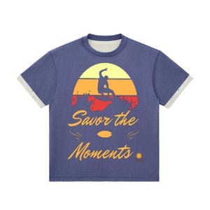 May include: Blue t-shirt with a surfer graphic. The graphic features a surfer silhouette riding a wave against a sunset backdrop with the text "Savor the Moments". The shirt has a crew neck and contrasting light grey sleeve cuffs.