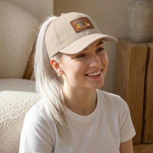 May include: A tan baseball cap with a brown leather patch featuring a cartoon design. The cap has a curved brim and a ponytail opening. The patch has yellow, brown, and white details. The cap is worn by a person with blonde hair.