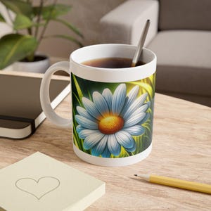 May include: White ceramic mug featuring a large, detailed daisy with blue petals and a yellow center. The mug is filled with a dark liquid and has a silver spoon inside. The design is set against a green and yellow background.