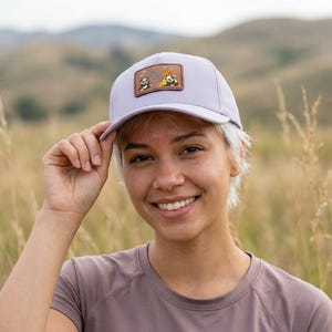 May include: A light purple baseball cap with a brown patch featuring panda illustrations and text. The cap is made of a soft material and has a curved brim. The background is a field of tall grass.