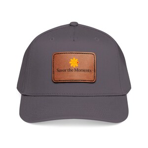 May include: A gray baseball cap with a curved brim. A brown leather patch on the front features a yellow flower with a red dot and the words "Savor the Moments" in a serif font.