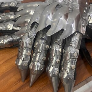 Puede incluir: Un par de guanteletes de metal articulado con placas de acero pulido en tono plateado y grabados intrincados. La armadura presenta puntas de dedos afiladas, placas de nudillos superpuestas y peque&ntilde;os remaches que aseguran los segmentos.
