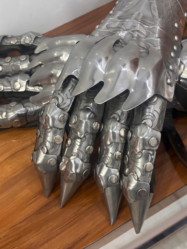 Puede incluir: Un par de guanteletes de metal articulado con placas de acero pulido en tono plateado y grabados intrincados. La armadura presenta puntas de dedos afiladas, placas de nudillos superpuestas y peque&ntilde;os remaches que aseguran los segmentos.