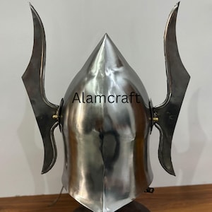 K&ouml;nnte beinhalten: Ein polierter Metallhelm mit spitzer Spitze und zwei gebogenen, fl&uuml;gelartigen Verl&auml;ngerungen. Der Helm ist auf einem Holzsockel montiert. Der Text "Alamcraft" ist auf dem Helm sichtbar.