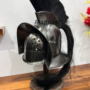 Könnte beinhalten: Ein römischer Gladiatorenhelm aus poliertem Metall mit schwarzer Feder und Wangenschutz. Der Helm wird auf einem Holzständer ausgestellt. Der Text "Alamcraft" ist auf der Seite des Helms zu sehen.