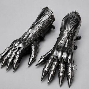 Puede incluir: Un par de guanteletes de metal articulado con intrincados patrones de volutas grabados, placas de dedos puntiagudas y un acabado plateado pulido. Las piezas de armadura incluyen un protector de antebrazo y el logo AC ALAH CRAFT a la derecha.