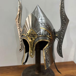 K&ouml;nnte beinhalten: Ein silberfarbener Helm mit goldenen Akzenten und dekorativen Seitenteilen. Der Helm hat eine spitze Spitze und wird auf einem Holzst&auml;nder pr&auml;sentiert. Der Text "Alamcraft" ist auf der Vorderseite sichtbar.