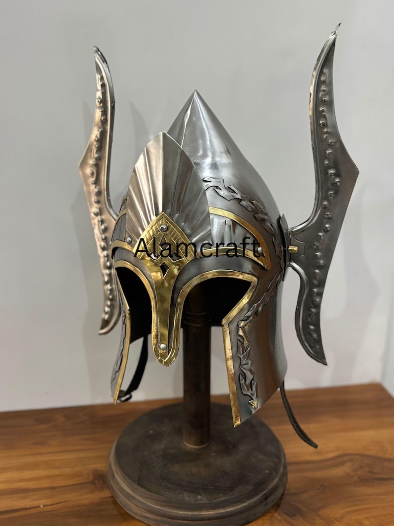 K&ouml;nnte beinhalten: Ein silberfarbener Helm mit goldenen Akzenten und dekorativen Seitenteilen. Der Helm hat eine spitze Spitze und wird auf einem Holzst&auml;nder pr&auml;sentiert. Der Text "Alamcraft" ist auf der Vorderseite sichtbar.