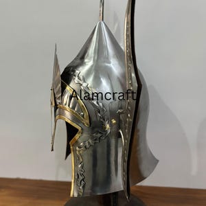 K&ouml;nnte beinhalten: Ein silberner Helm mit goldenen Akzenten und aufwendigen Blattmustern. Der Helm hat eine spitze Spitze und l&auml;ngliche Seitenteile. Der Text "Alamcraft" ist auf der Seite sichtbar.