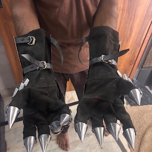 Puede incluir: Un par de guantes tipo guantelete de ante marr&oacute;n oscuro con largas garras met&aacute;licas plateadas y puntiagudas en cada dedo. Los guantes incluyen correas de cuero negro ajustables con hebillas met&aacute;licas alrededor de las mu&ntilde;ecas y antebrazos.