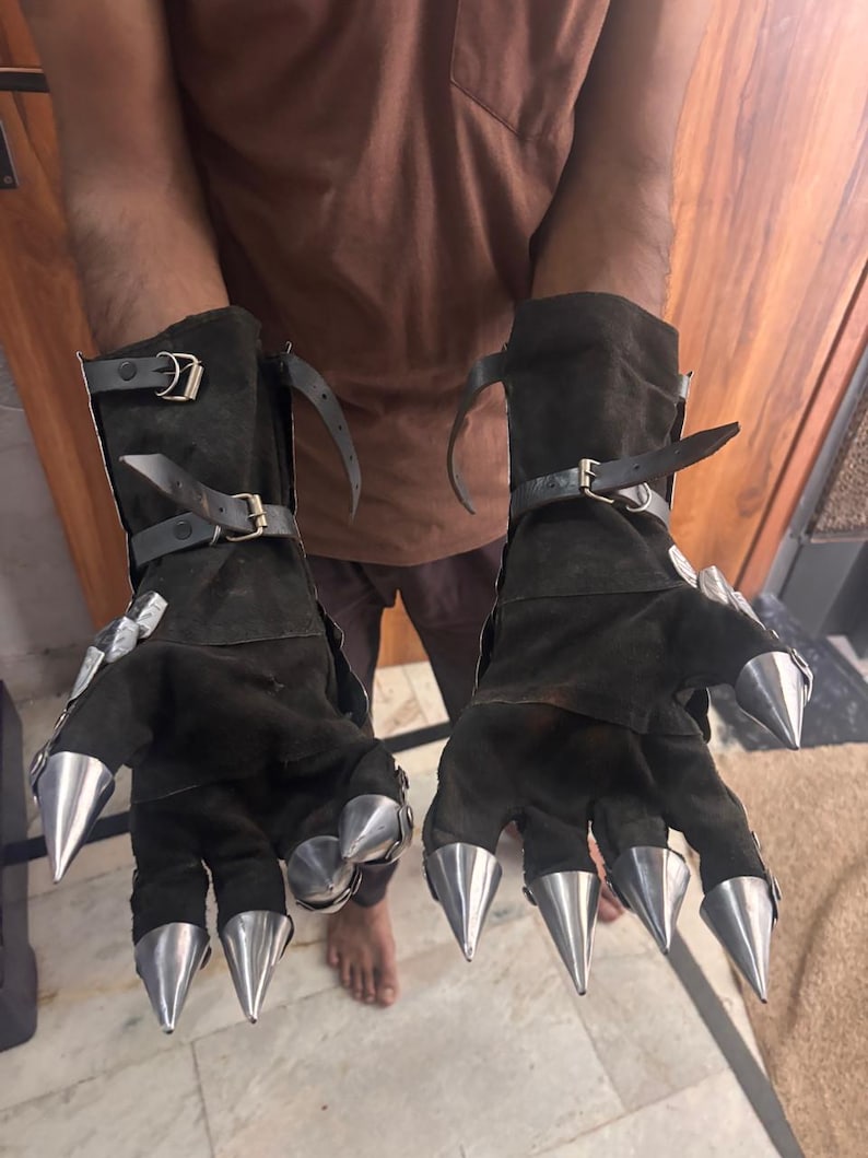 Puede incluir: Un par de guantes tipo guantelete de ante marr&oacute;n oscuro con largas garras met&aacute;licas plateadas y puntiagudas en cada dedo. Los guantes incluyen correas de cuero negro ajustables con hebillas met&aacute;licas alrededor de las mu&ntilde;ecas y antebrazos.