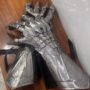 Puede incluir: Un guantelete de metal plateado con placas articuladas para los dedos con puntas afiladas, intrincados patrones de volutas en relieve y una secci&oacute;n de antebrazo acampanada. La pieza de armadura incluye correas de cuero negro para sujetar al brazo.