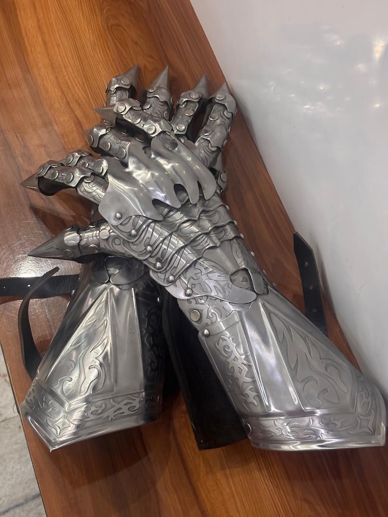 Puede incluir: Un guantelete de metal plateado con placas articuladas para los dedos con puntas afiladas, intrincados patrones de volutas en relieve y una secci&oacute;n de antebrazo acampanada. La pieza de armadura incluye correas de cuero negro para sujetar al brazo.