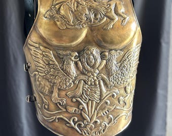 May include: A gold-colored breastplate with intricate relief carvings. The design features mythical creatures, including lions and winged figures, and ornate floral patterns. The breastplate has a polished finish and is displayed against a dark background.