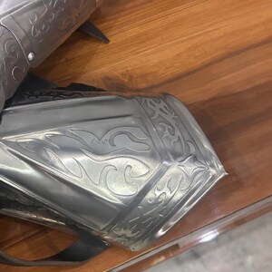Puede incluir: Un par de guanteletes de metal color plata pulida con intrincados grabados en relieve de volutas y patrones de llamas. Las piezas de armadura est&aacute;n construidas con placas articuladas e incluyen correas de cuero negro visibles para sujeci&oacute;n.