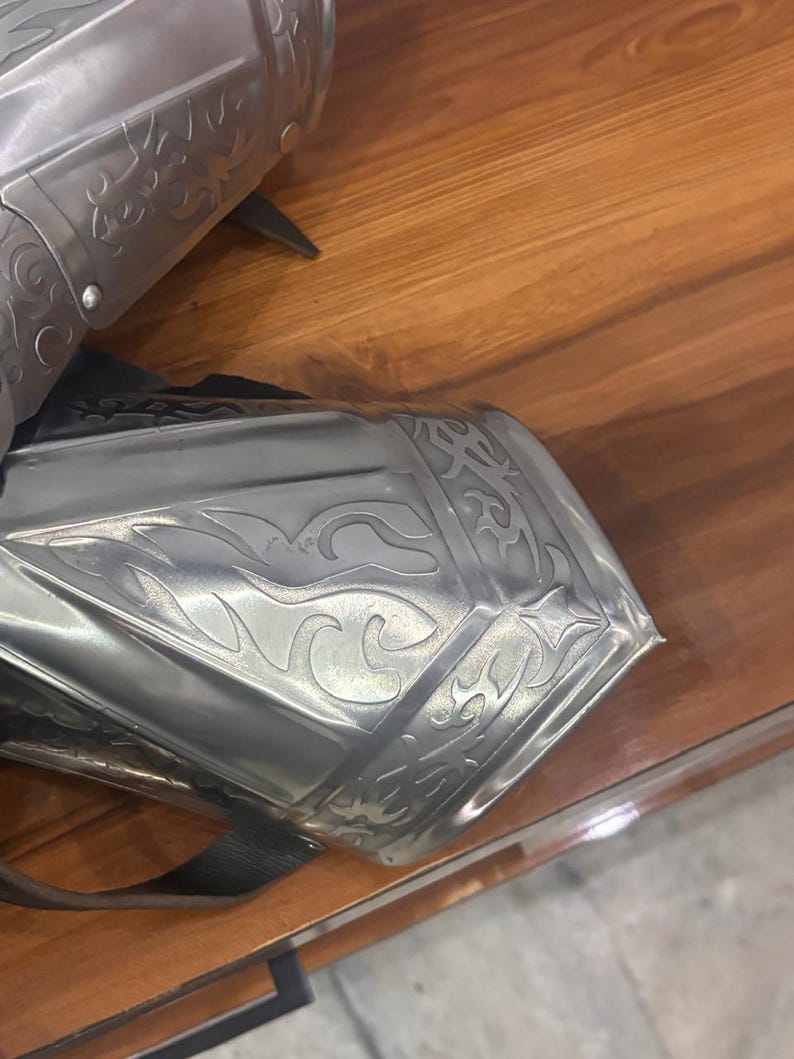 Puede incluir: Un par de guanteletes de metal color plata pulida con intrincados grabados en relieve de volutas y patrones de llamas. Las piezas de armadura est&aacute;n construidas con placas articuladas e incluyen correas de cuero negro visibles para sujeci&oacute;n.