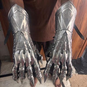 Puede incluir: Un par de guanteletes de metal plateado pulido con intrincados patrones de volutas grabados en las placas del antebrazo. Las secciones articuladas de los dedos terminan en puntas afiladas como garras y est&aacute;n aseguradas con remaches y correas de cuero negro.