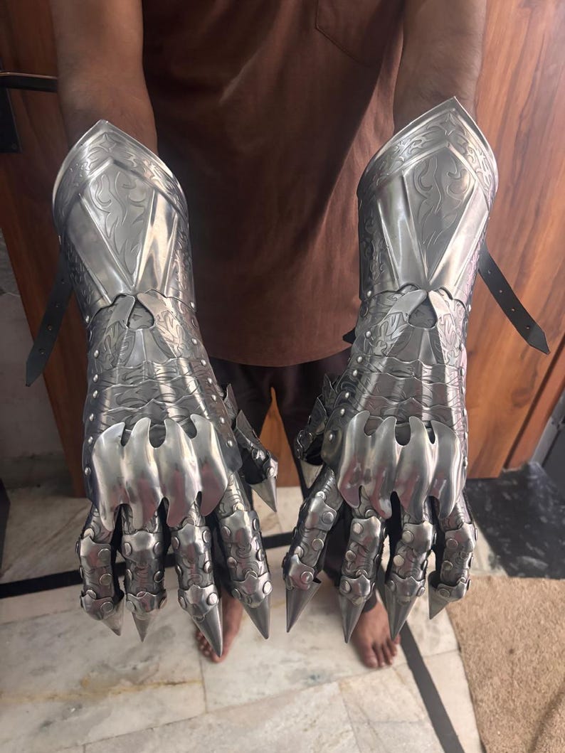 Puede incluir: Un par de guanteletes de metal plateado pulido con intrincados patrones de volutas grabados en las placas del antebrazo. Las secciones articuladas de los dedos terminan en puntas afiladas como garras y est&aacute;n aseguradas con remaches y correas de cuero negro.