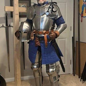 Könnte beinhalten: Eine vollständige Rüstung aus poliertem Stahlblech mit Brustpanzer, Schulterstücken und Beinschienen, getragen über einem blauen, gesteppten Gambeson. Die Rüstung umfasst einen passenden Stahlhelm in einer Hand, einen braunen Ledergürtel mit Tasche und ein Schwert an der Hüfte. Das Logo AALAM CRAFT ist auf dem Brustpanzer sichtbar.