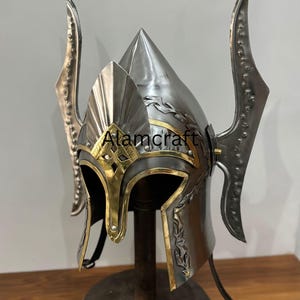 K&ouml;nnte beinhalten: Ein silberner Helm mit goldenen Akzenten und dekorativen Elementen. Der Helm hat eine spitze Spitze und gebogene Seitenteile. Der Text "Alamcraft" ist sichtbar. Er wird auf einem Holzst&auml;nder pr&auml;sentiert.