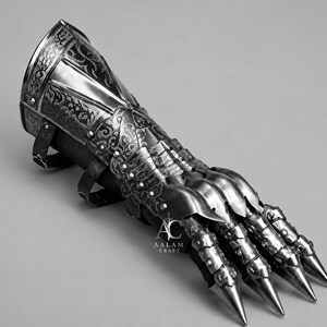 Puede incluir: Un guantelete de acero pulido con intrincados patrones florales grabados, placas articuladas para los dedos y puntas afiladas. La armadura de metal incluye correas de cuero negro para sujetar y muestra el texto AALAM CRAFT con un icono de espada.