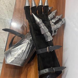 Puede incluir: Un par de guanteletes negros con placas de dedos articuladas de metal plateado y puntas afiladas. Los guanteletes incluyen un protector de antebrazo met&aacute;lico con grabados intrincados y dos correas de cuero negro ajustables con hebillas.