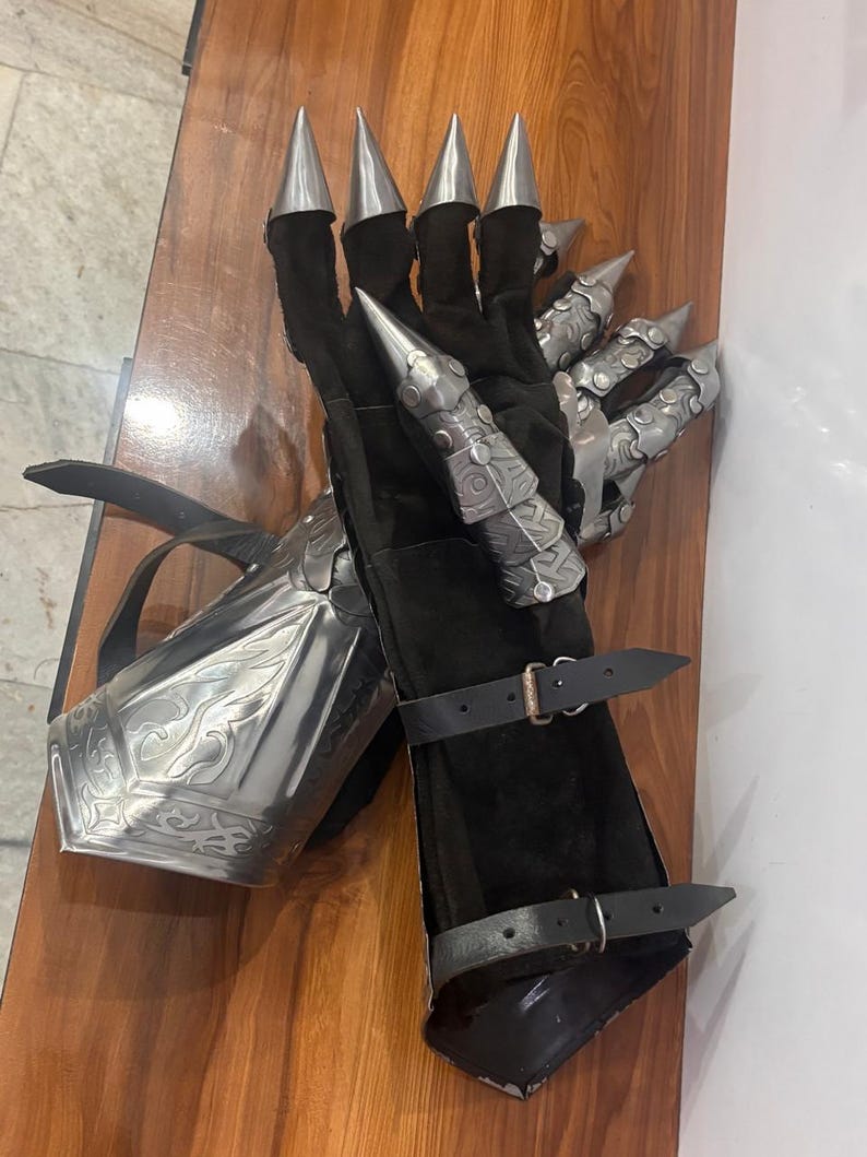 Puede incluir: Un par de guanteletes negros con placas de dedos articuladas de metal plateado y puntas afiladas. Los guanteletes incluyen un protector de antebrazo met&aacute;lico con grabados intrincados y dos correas de cuero negro ajustables con hebillas.