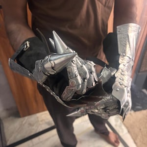 Puede incluir: Un par de guanteletes de metal articulado con puntas en los dedos y complejos patrones de volutas grabados. Los guanteletes est&aacute;n construidos con placas de tono plateado pulido sobre una base de tela oscura, asegurados con correas ajustables.