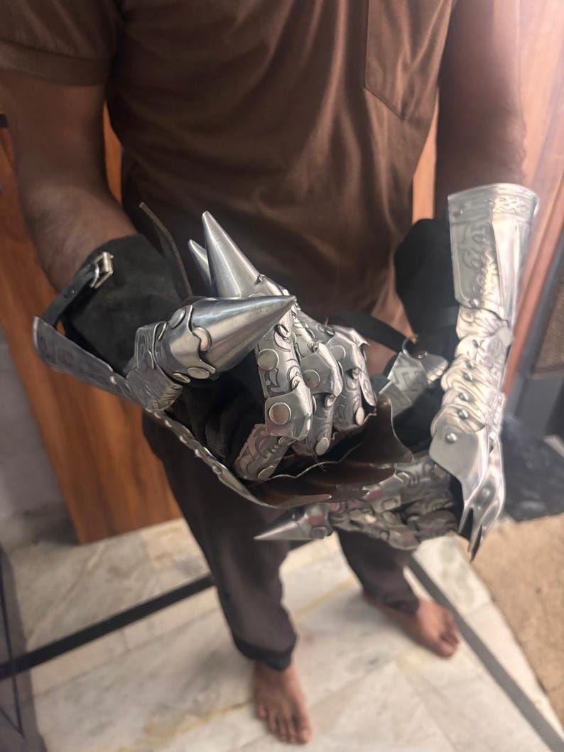 Puede incluir: Un par de guanteletes de metal articulado con puntas en los dedos y complejos patrones de volutas grabados. Los guanteletes est&aacute;n construidos con placas de tono plateado pulido sobre una base de tela oscura, asegurados con correas ajustables.