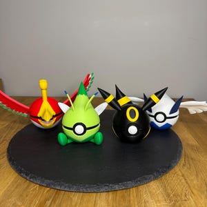 Könnte beinhalten: Vier farbenfrohe, stilisierte Figuren von Pokémon-Charakteren sind auf einer schwarzen, kreisförmigen Oberfläche angeordnet. Die Figuren umfassen eine rot-gelbe vogelartige Figur, eine grüne pflanzenartige Figur, eine schwarze Figur mit gelben Akzenten und eine blau-weiße Figur.